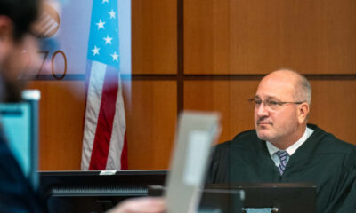 A judge in a black robe sits at a courtroom bench, with an American flag in the background.