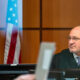 A judge in a black robe sits at a courtroom bench, with an American flag in the background.