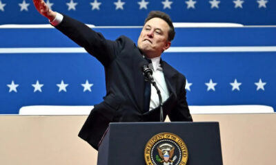 Man in a dark suit at a podium with the presidential seal, raising one arm in front of a blue starry backdrop.