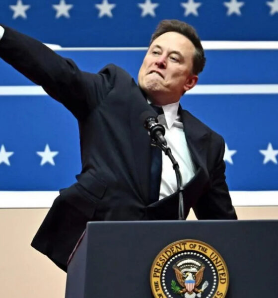 Man in a dark suit at a podium with the presidential seal, raising one arm in front of a blue starry backdrop.