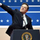Man in a dark suit at a podium with the presidential seal, raising one arm in front of a blue starry backdrop.