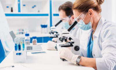 Two researchers in lab coats, masks, and gloves examine samples through compound microscopes at a white benchtop lab setup with blue accents.