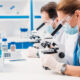 Two researchers in lab coats, masks, and gloves examine samples through compound microscopes at a white benchtop lab setup with blue accents.