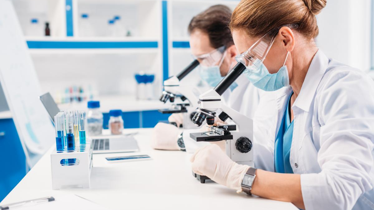 Two researchers in lab coats, masks, and gloves examine samples through compound microscopes at a white benchtop lab setup with blue accents.