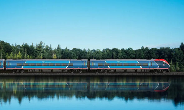 Blue Amtrak passenger train traveling on tracks beside a calm lake with trees in the background and a clear sky, reflected in the water.