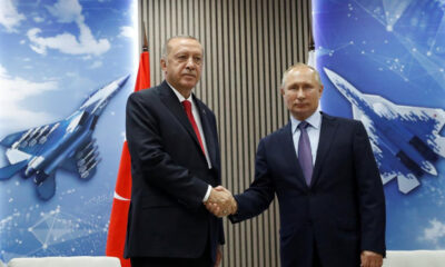 Two men in dark suits shake hands in front of a blue, aerospace-themed backdrop and a red national flag behind them.