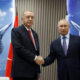 Two men in dark suits shake hands in front of a blue, aerospace-themed backdrop and a red national flag behind them.