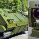 Green armored tracked vehicle beside a large gyrostabilized optical sensor with purple lenses on the right side.