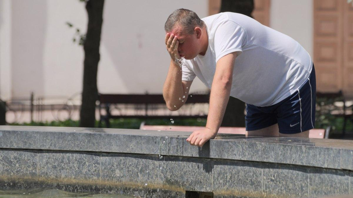 Man in a white T-shirt and navy shorts leans over a stone fountain, dabbing water from his face with his hand as water droplets fall.