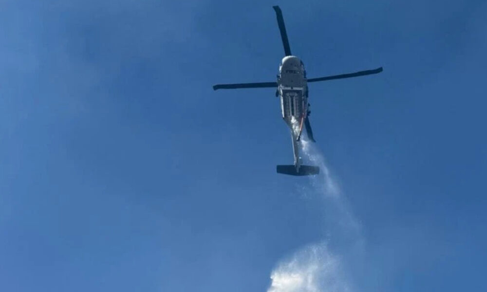 A firefighting helicopter releasing water over a clear blue sky, with a stream forming below the aircraft.