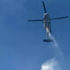 A firefighting helicopter releasing water over a clear blue sky, with a stream forming below the aircraft.