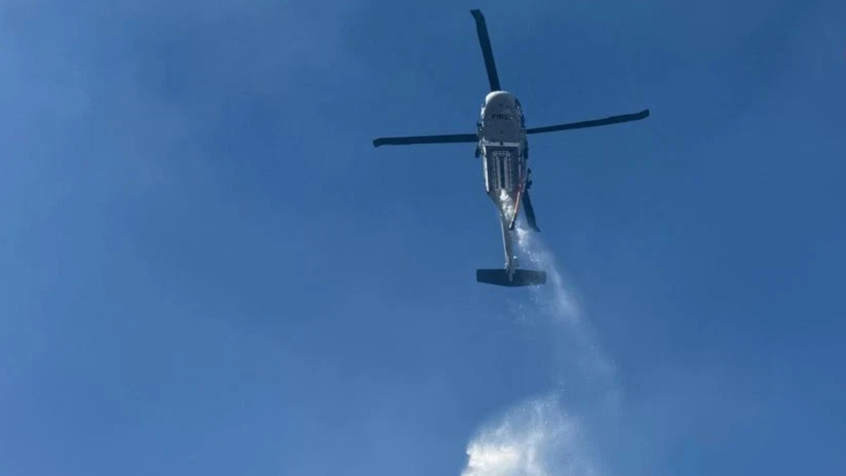 A firefighting helicopter releasing water over a clear blue sky, with a stream forming below the aircraft.