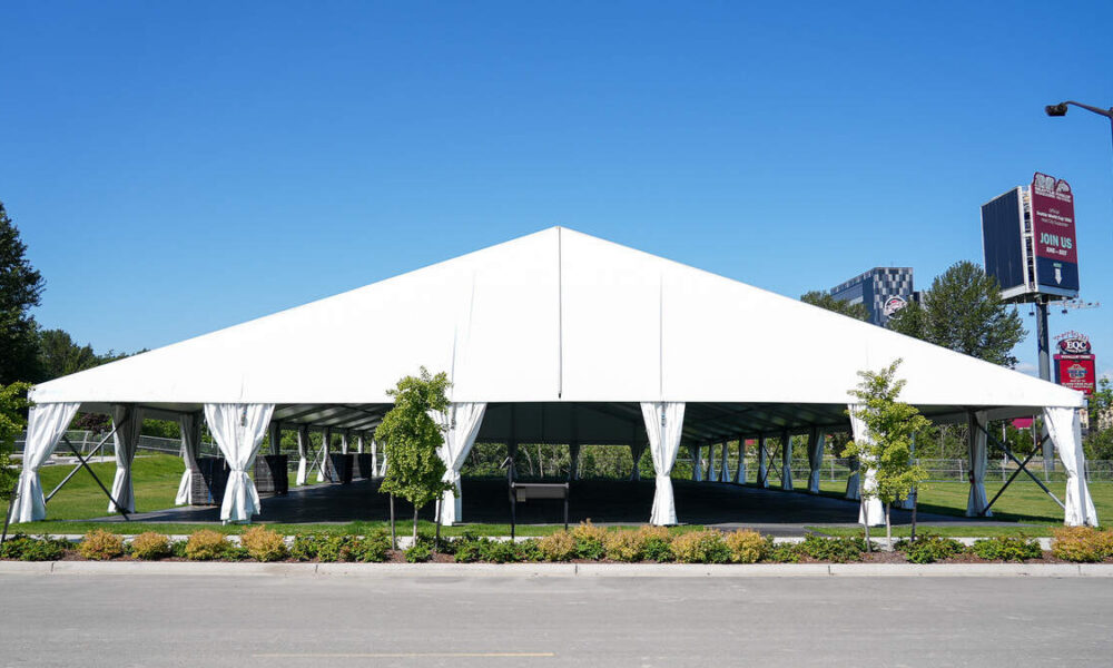 Large white outdoor event tent with open sides and tied-back curtains on a grassy lot, under a clear blue sky.