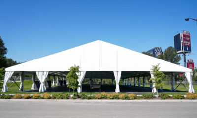 Large white outdoor event tent with open sides and tied-back curtains on a grassy lot, under a clear blue sky.