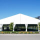 Large white outdoor event tent with open sides and tied-back curtains on a grassy lot, under a clear blue sky.