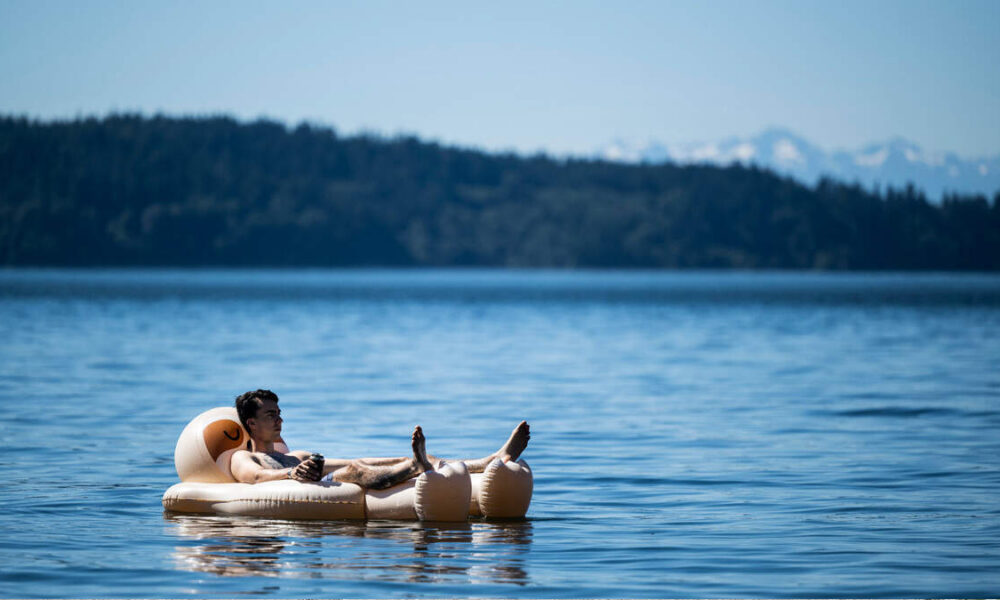 Man relaxing on a beige inflatable raft on a calm blue lake, with forested hills and distant mountains in the background.