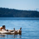 Man relaxing on a beige inflatable raft on a calm blue lake, with forested hills and distant mountains in the background.