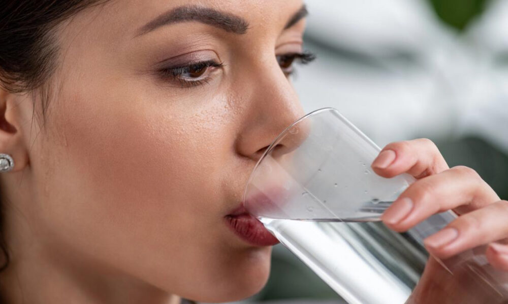 Close-up of a woman sipping water from a clear glass, with a neutral expression and an earring visible on her ear.
