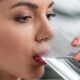 Close-up of a woman sipping water from a clear glass, with a neutral expression and an earring visible on her ear.
