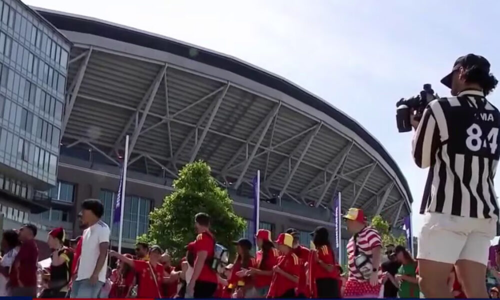Group of people in red outfits lined up outside a curved stadium, with a photographer in a black-and-white striped shirt filming the scene.
