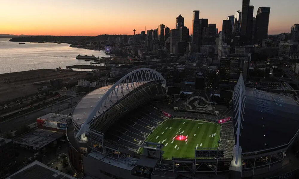 Aerial view of a large football stadium by the water at sunset, with a city skyline in the background.