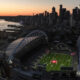 Aerial view of a large football stadium by the water at sunset, with a city skyline in the background.