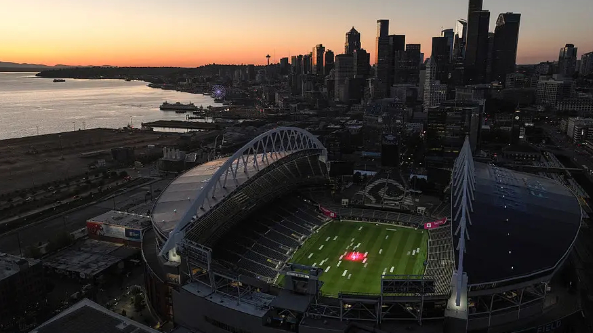 Aerial view of a large football stadium by the water at sunset, with a city skyline in the background.