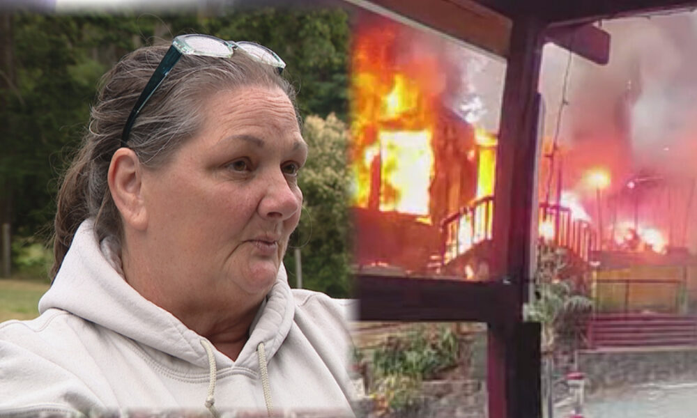 Woman in a light hoodie with glasses on her head is being interviewed outdoors, as a burning building smolders in the background.