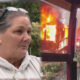 Woman in a light hoodie with glasses on her head is being interviewed outdoors, as a burning building smolders in the background.