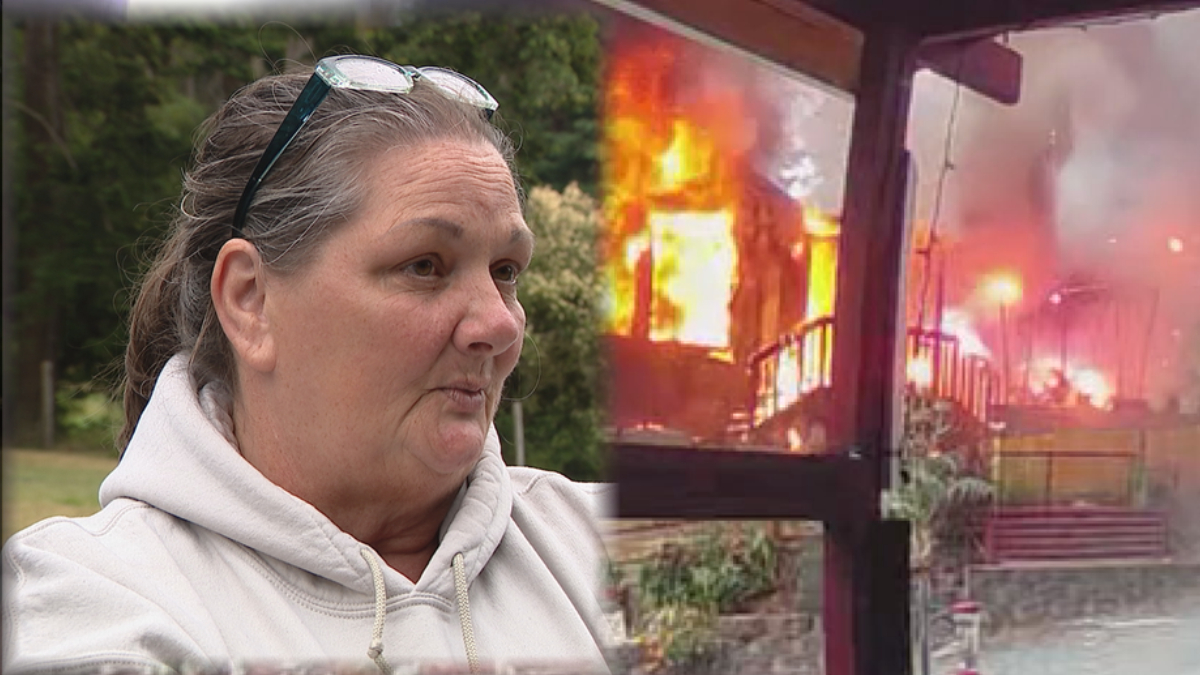 Woman in a light hoodie with glasses on her head is being interviewed outdoors, as a burning building smolders in the background.