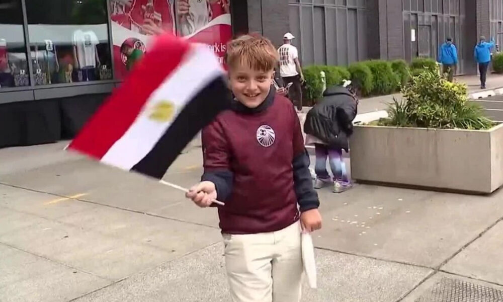 Smiling boy in a maroon jacket waves an Egyptian flag on a city sidewalk with storefronts in the background.
