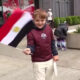 Smiling boy in a maroon jacket waves an Egyptian flag on a city sidewalk with storefronts in the background.