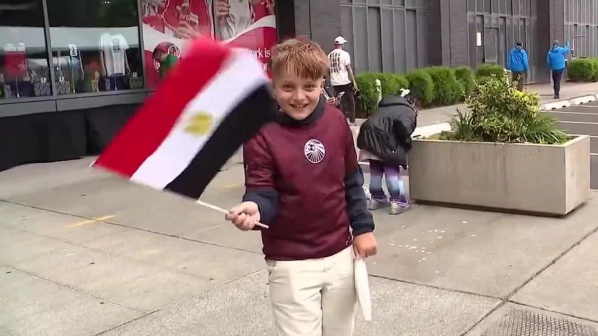 Smiling boy in a maroon jacket waves an Egyptian flag on a city sidewalk with storefronts in the background.