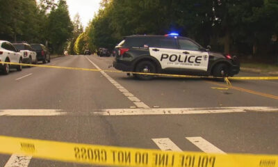 Police SUV with blue lights blocking a tree-lined street, yellow caution tape stretched across the road in a residential area.