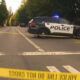 Police SUV with blue lights blocking a tree-lined street, yellow caution tape stretched across the road in a residential area.