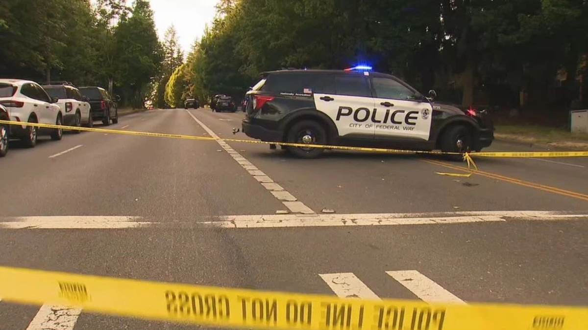 Police SUV with blue lights blocking a tree-lined street, yellow caution tape stretched across the road in a residential area.