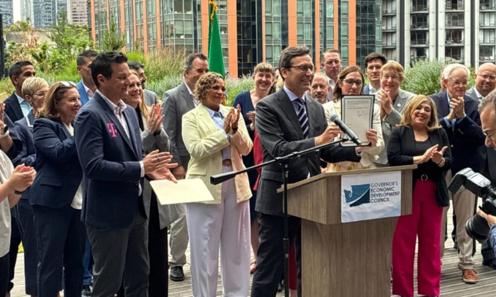 Man in a suit holding a framed certificate at a podium as a diverse group applauds at an outdoor event with a Governor's Economic Development Council banner on the podium.