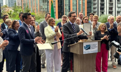 Man in a suit holding a framed certificate at a podium as a diverse group applauds at an outdoor event with a Governor's Economic Development Council banner on the podium.