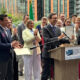 Man in a suit holding a framed certificate at a podium as a diverse group applauds at an outdoor event with a Governor's Economic Development Council banner on the podium.