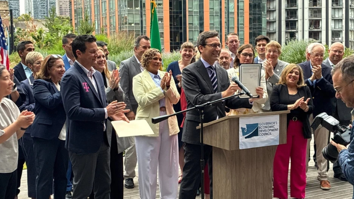 Man in a suit holding a framed certificate at a podium as a diverse group applauds at an outdoor event with a Governor's Economic Development Council banner on the podium.