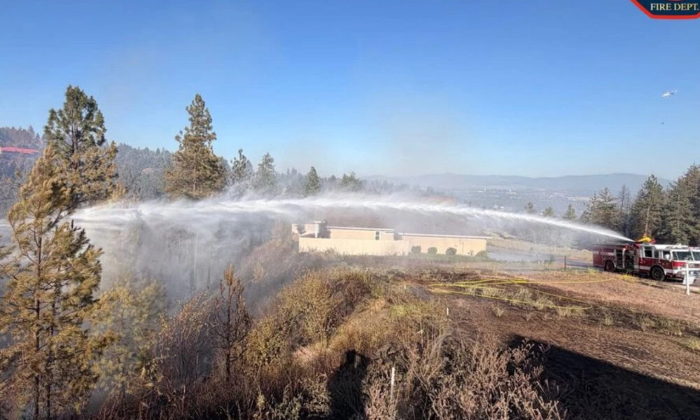 Firefighters on a hill spray water from two trucks to fight a brush fire near a beige building, against a blue sky.