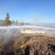 Firefighters on a hill spray water from two trucks to fight a brush fire near a beige building, against a blue sky.