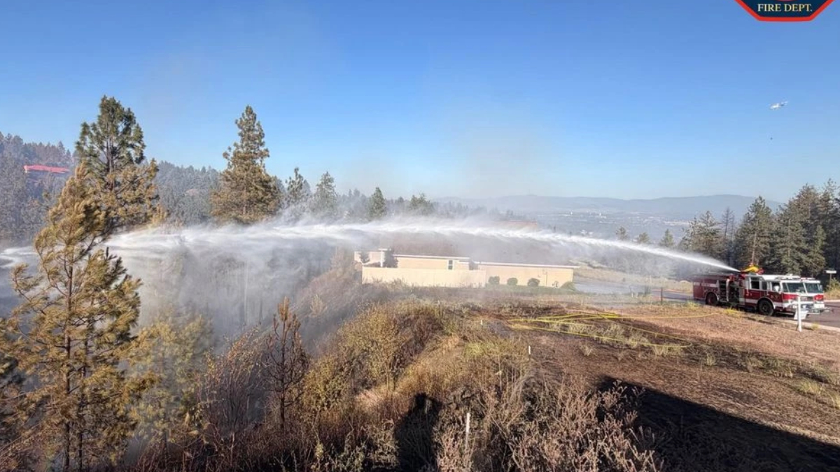 Firefighters on a hill spray water from two trucks to fight a brush fire near a beige building, against a blue sky.