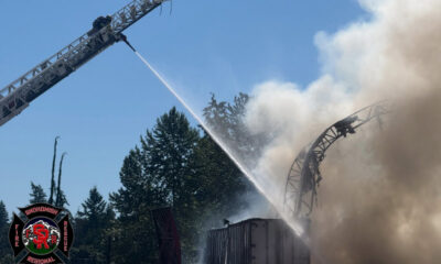 Firefighters on a ladder truck spray water on a burning structure, with heavy smoke and trees nearby.