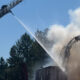 Firefighters on a ladder truck spray water on a burning structure, with heavy smoke and trees nearby.