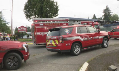 Emergency scene with red medic unit and fire truck on a street, motel sign in background, caution tape nearby.