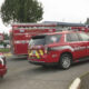 Emergency scene with red medic unit and fire truck on a street, motel sign in background, caution tape nearby.