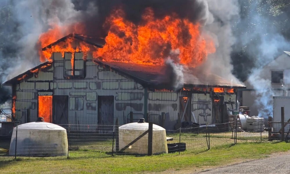House on fire with large flames and smoke, propane tanks in the foreground and a fenced yard nearby.