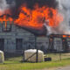 House on fire with large flames and smoke, propane tanks in the foreground and a fenced yard nearby.