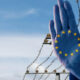 Blue protective glove with the EU circle of twelve yellow stars, raised against a barbed-wire border fence, symbolizing Europe's border control or solidarity.